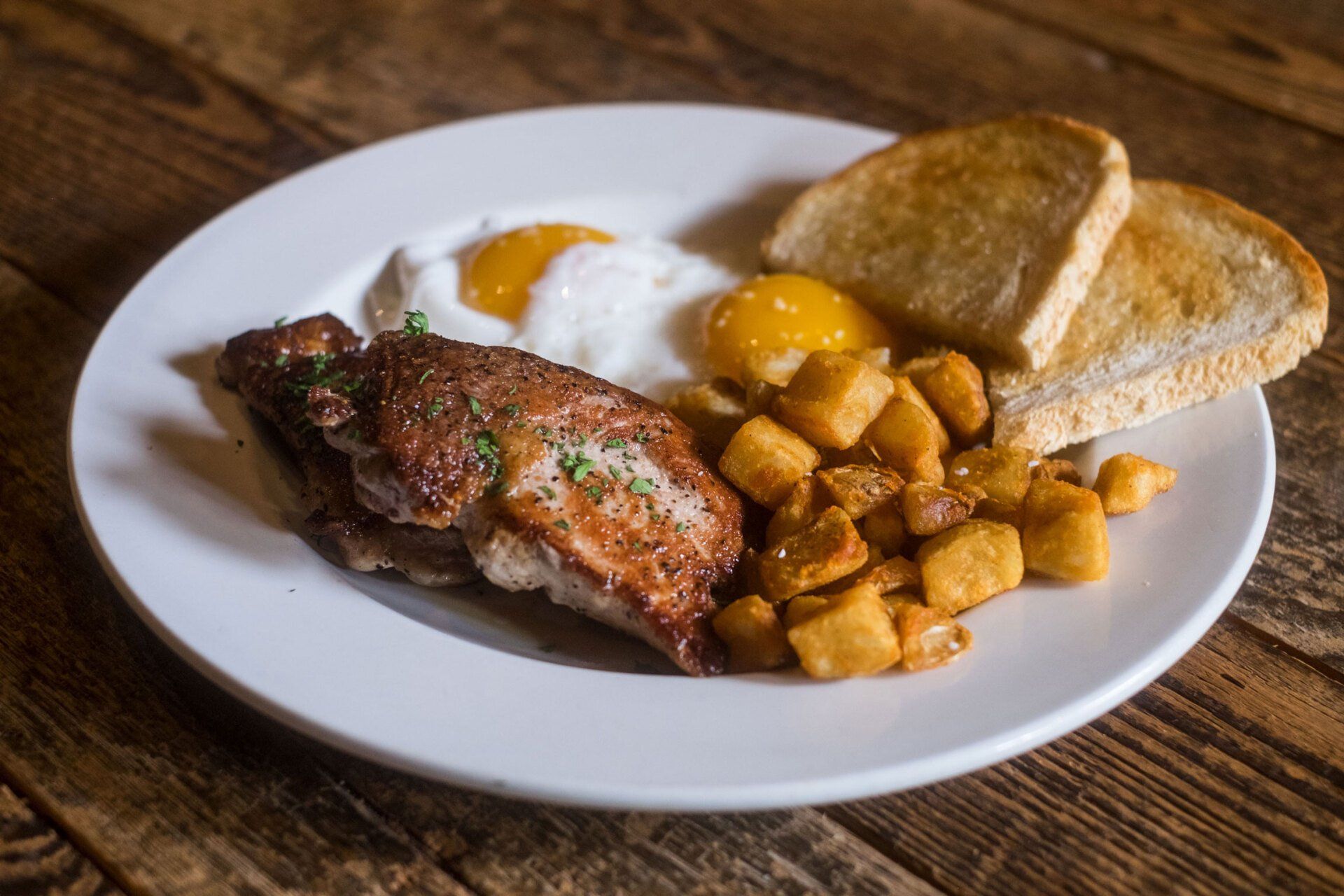 A white plate topped with meat , eggs , potatoes and toast on a wooden table.