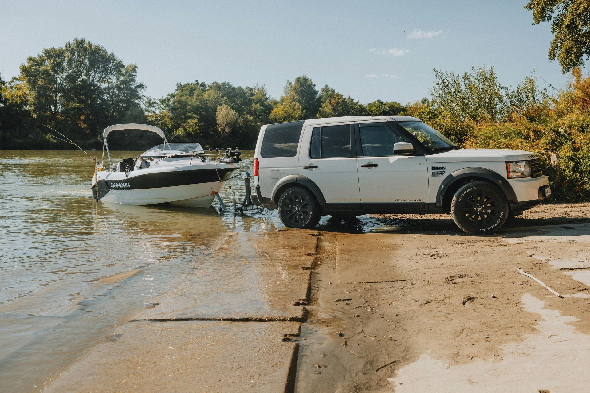 White SUV towing a boat into a body of water at a boat launch on a sunny day.