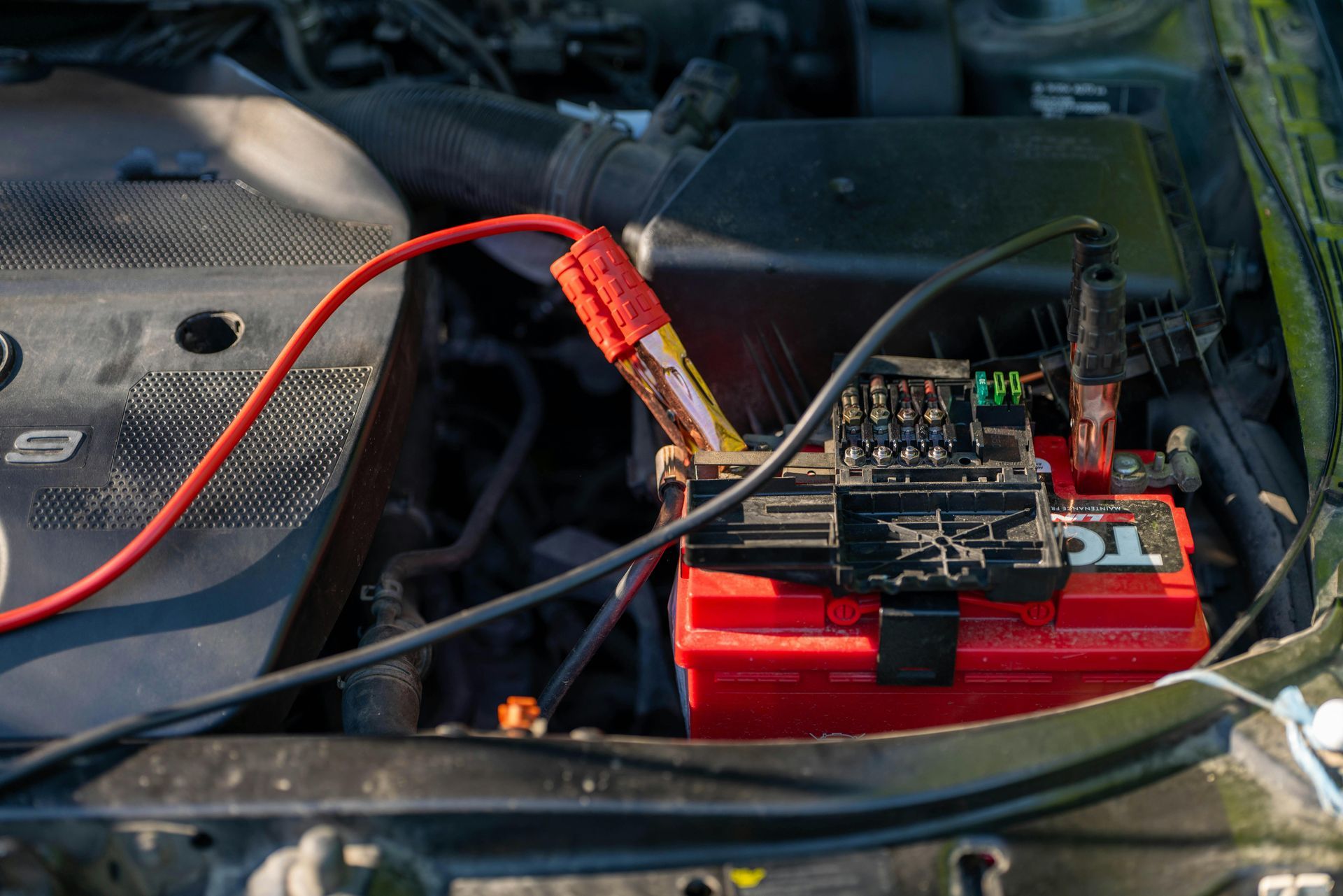 Car battery being jump-started; red and black jumper cables connected to a red battery in engine bay.