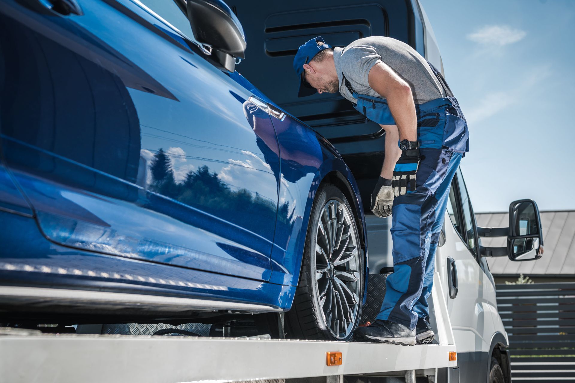 A tow truck operator secures a blue car onto a flatbed on a sunny day.