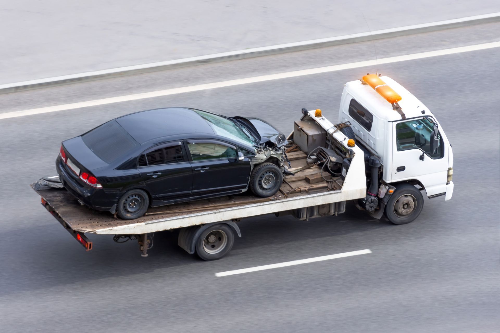 Black car damaged on a flatbed tow truck traveling on a highway.