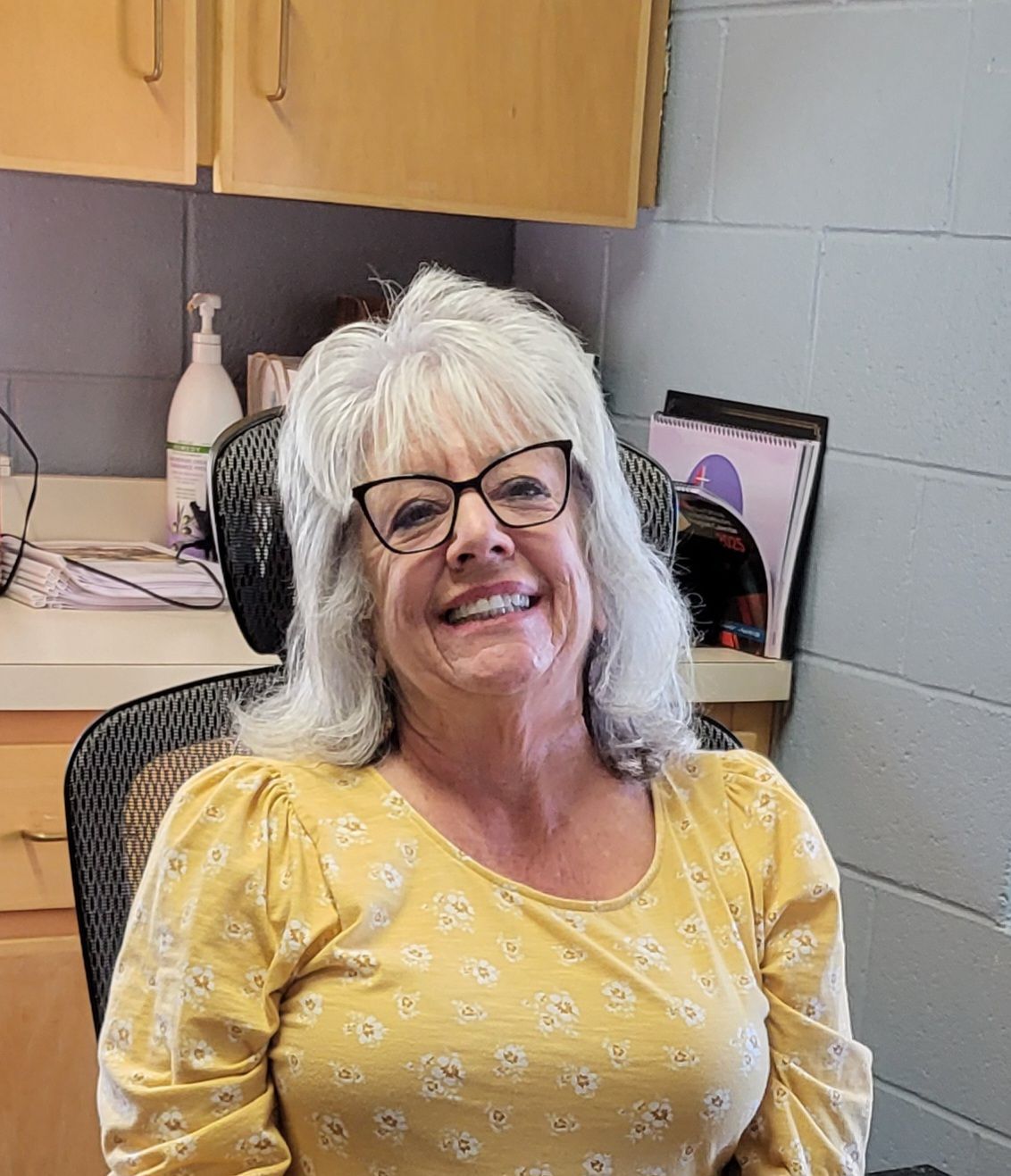Woman with gray hair, wearing glasses and yellow floral shirt, smiles in office setting.