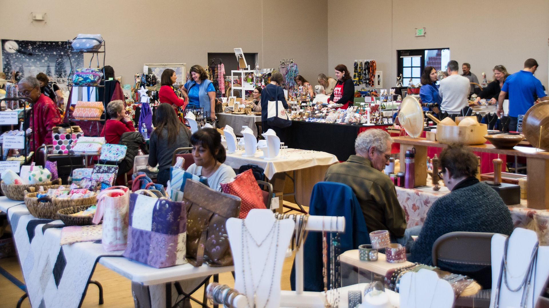 Craft fair with vendors and shoppers browsing tables filled with goods inside a large hall.