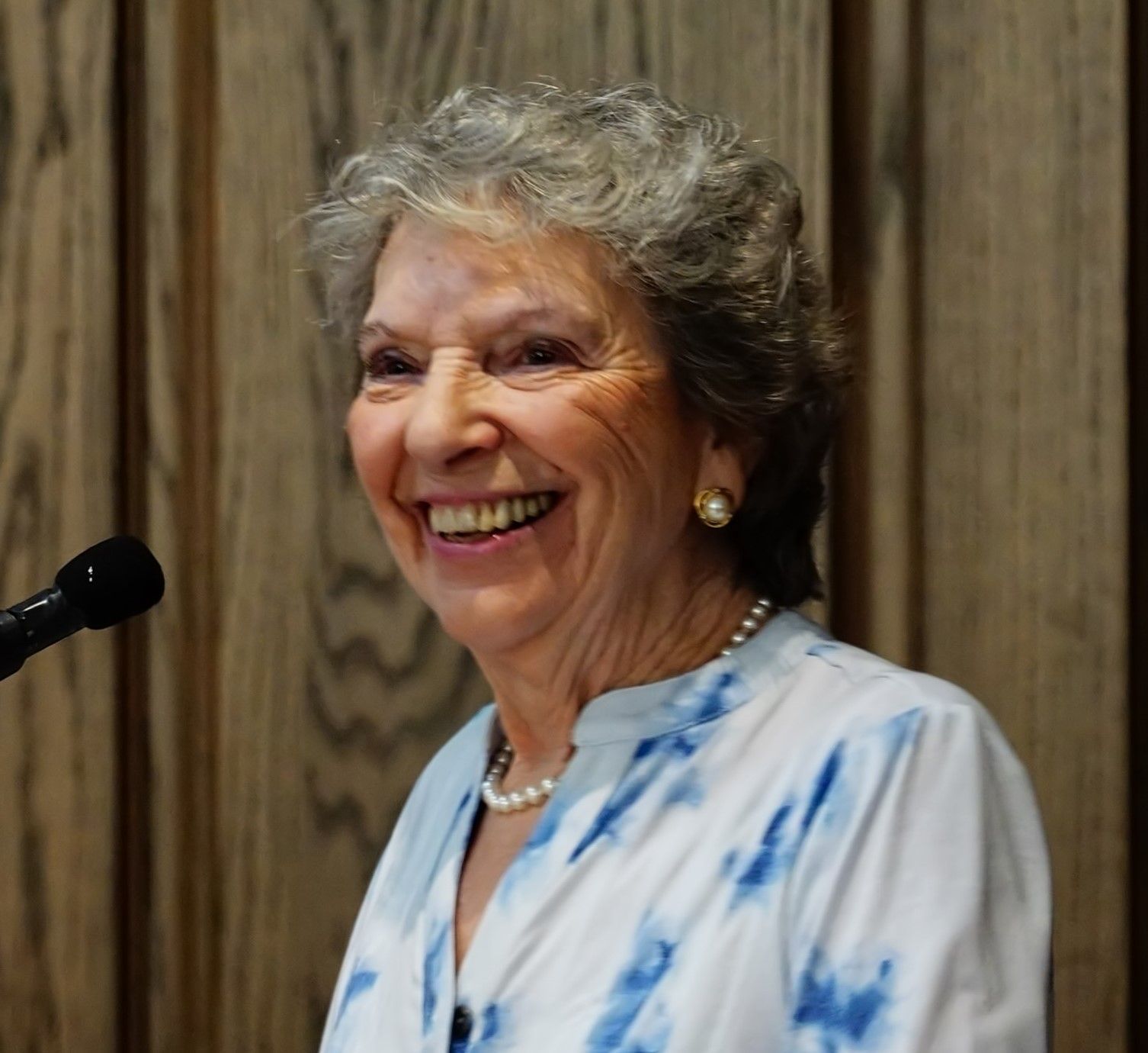 Woman smiling, speaking at a podium. Wearing a blue-and-white tie-dye shirt and pearl necklace.