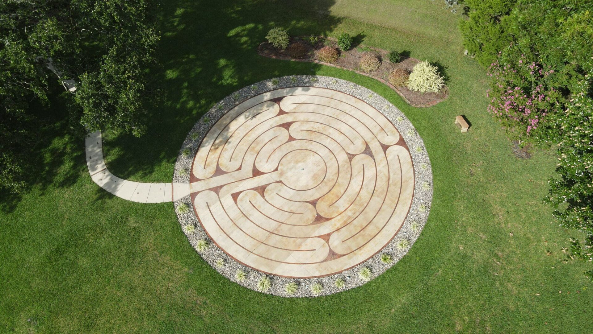 Overhead view of a circular stone labyrinth in a grassy yard, with a pathway leading to it.