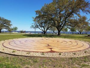 Circular labyrinth path in a park, surrounded by pebbles, grass, and trees. Clear blue sky.