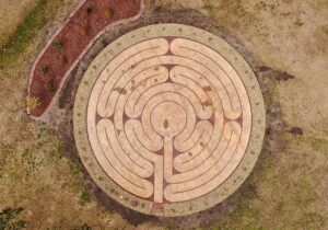Overhead view of a circular, beige labyrinth with a path leading to its center, surrounded by brown and green ground cover.