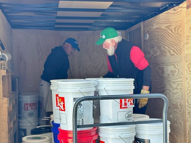 Two men inside a trailer, working with white buckets. One wears a green hat, the other a blue hat.