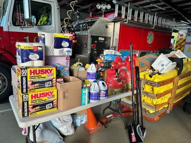 Supplies on a table in front of a fire truck: paper towels, cleaning supplies, tools, sandbags.