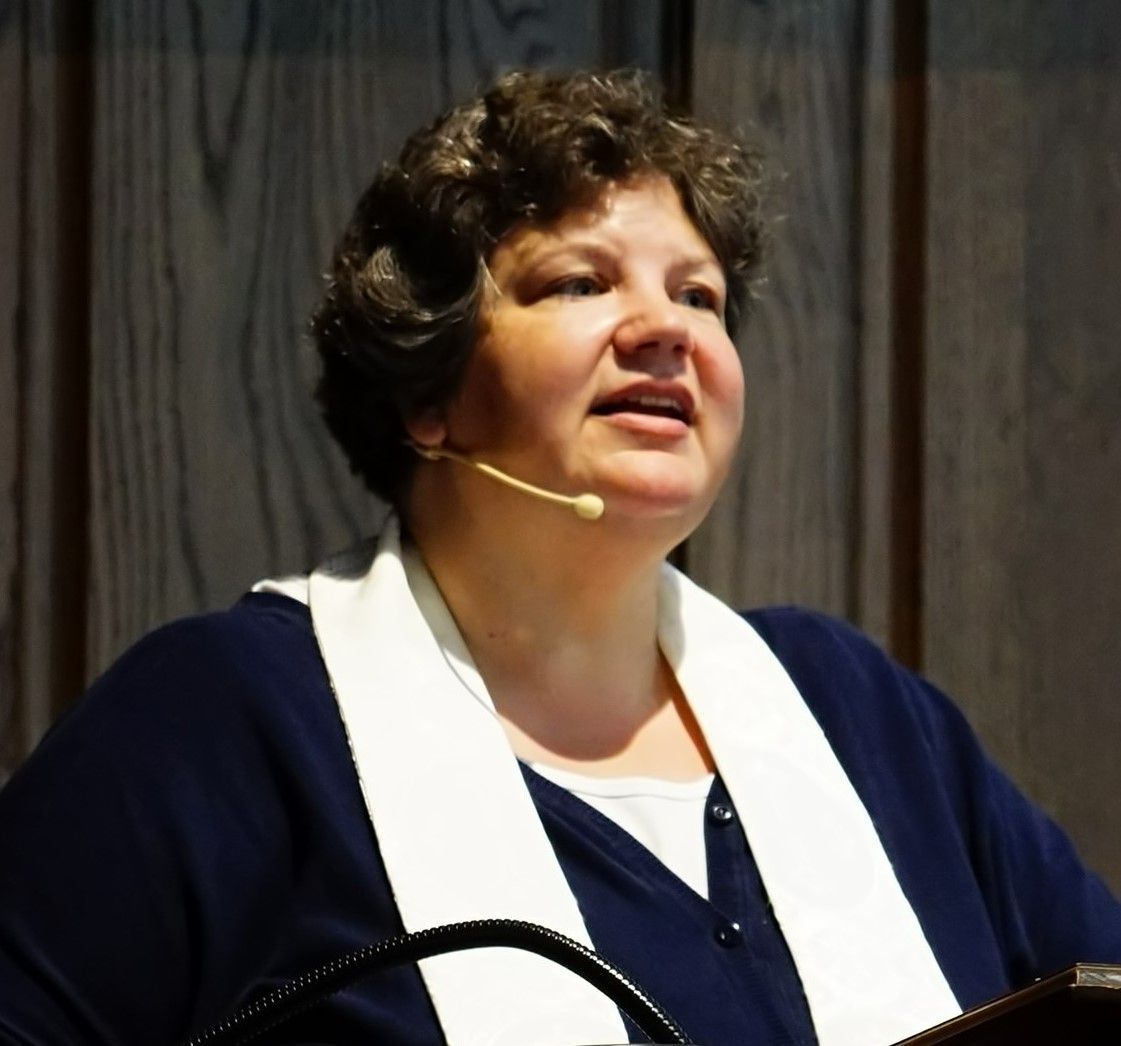 Woman speaking at a lectern, wearing a dark blue sweater and white collar; microphone near her mouth.