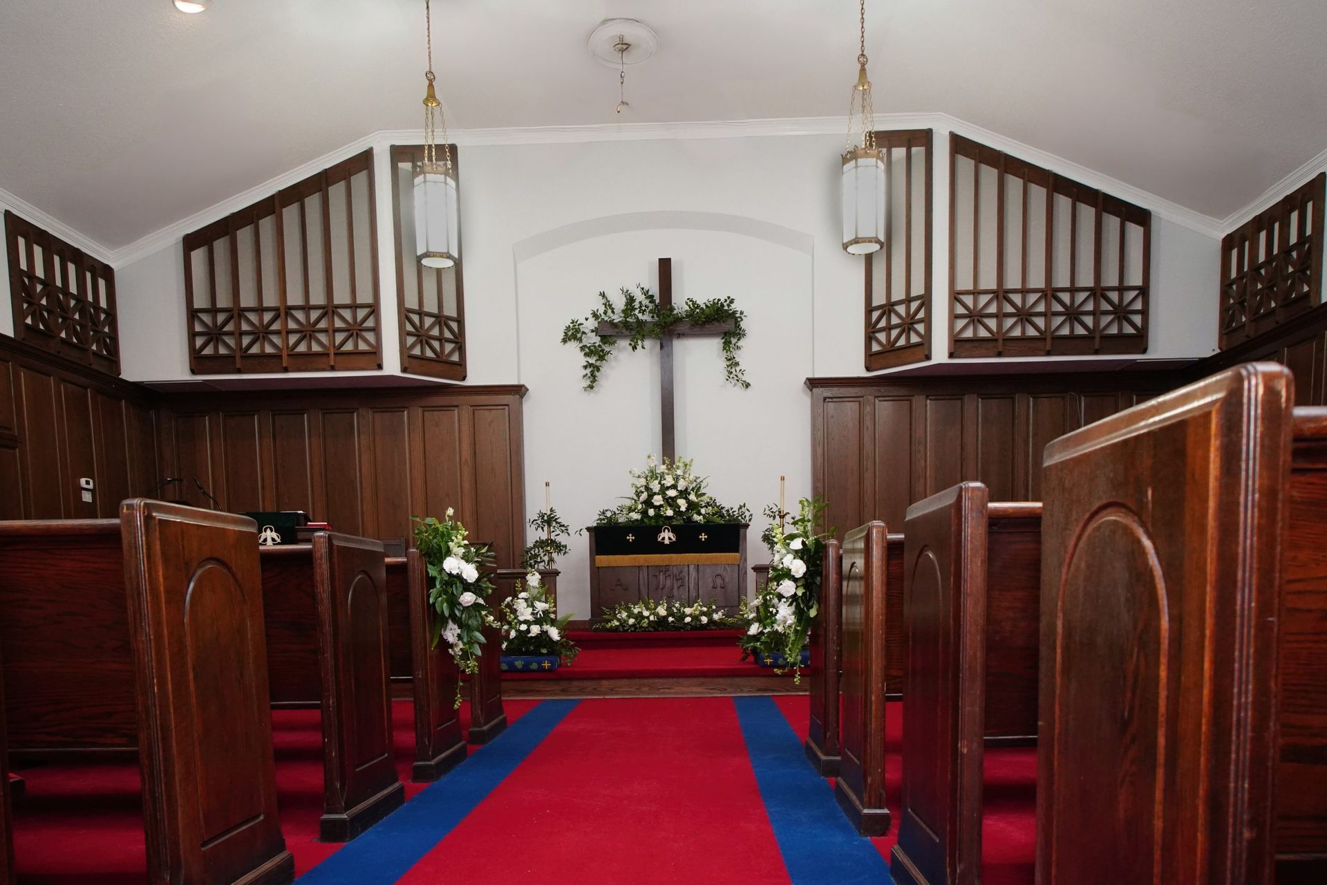 Interior of a chapel with wooden pews, red carpet, and a cross decorated with flowers.