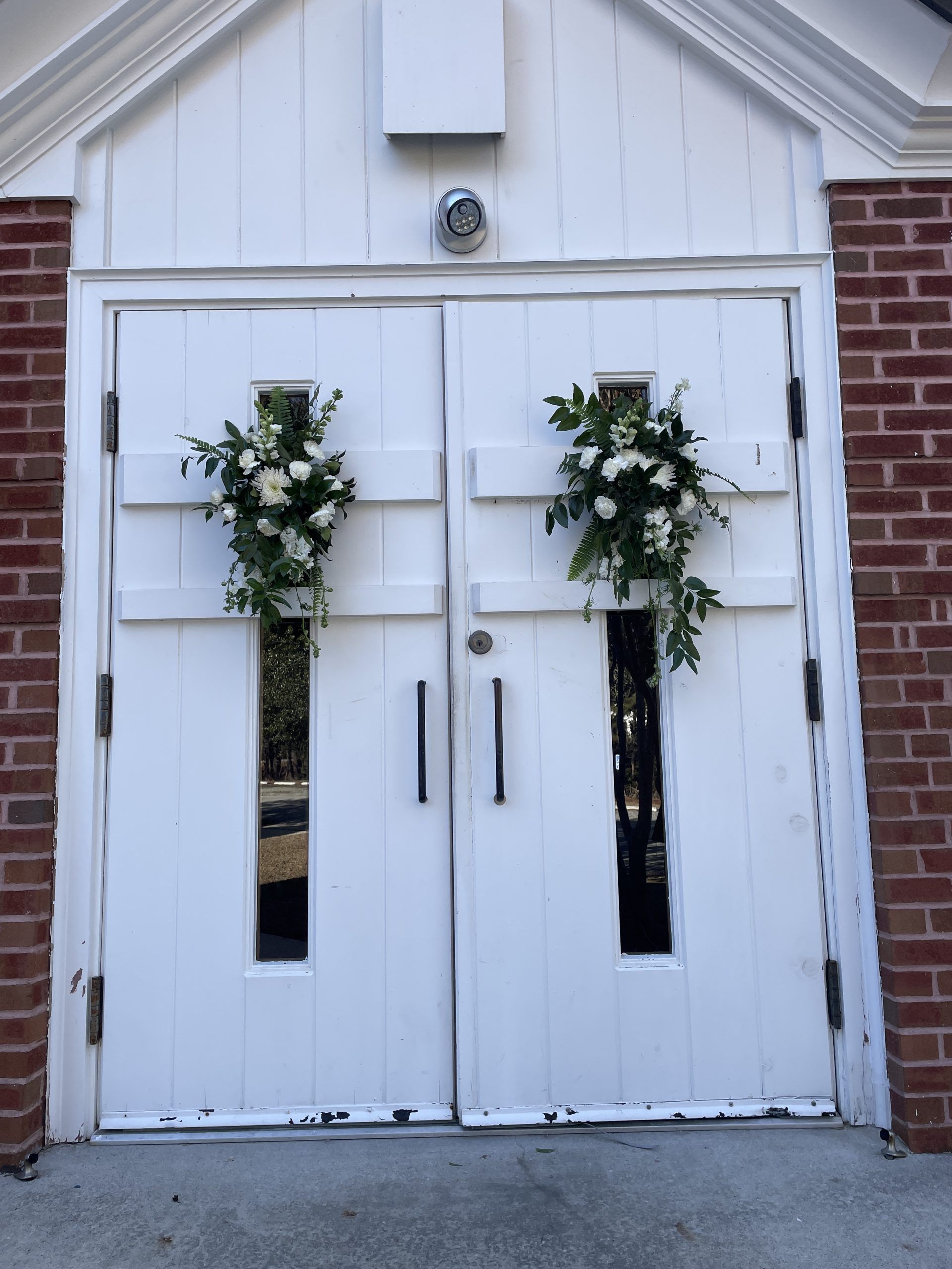White double doors decorated with floral arrangements, framed by brick.
