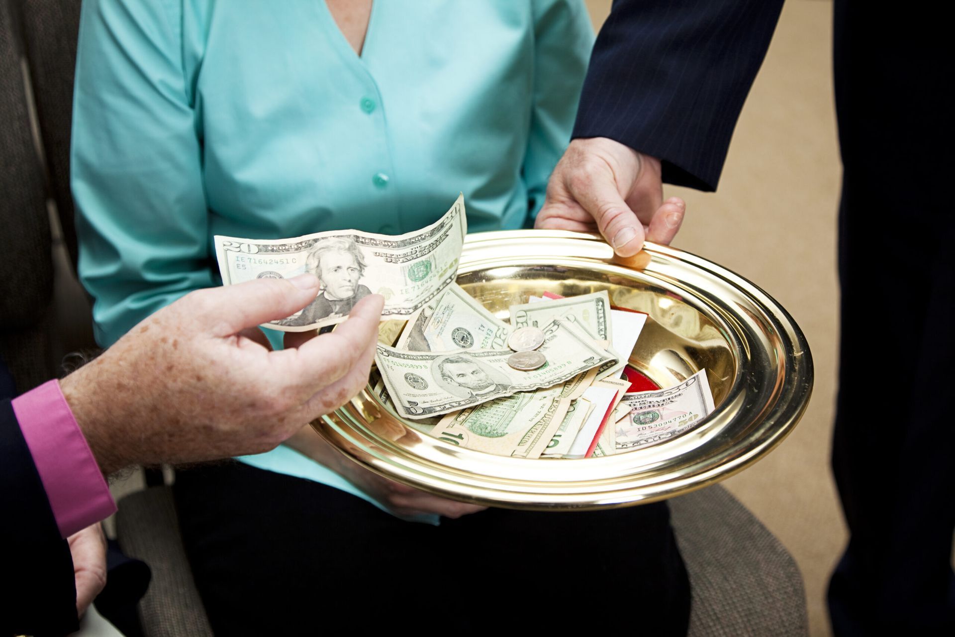 Hands holding a gold plate with cash during an offering. A person is handing money onto the plate.