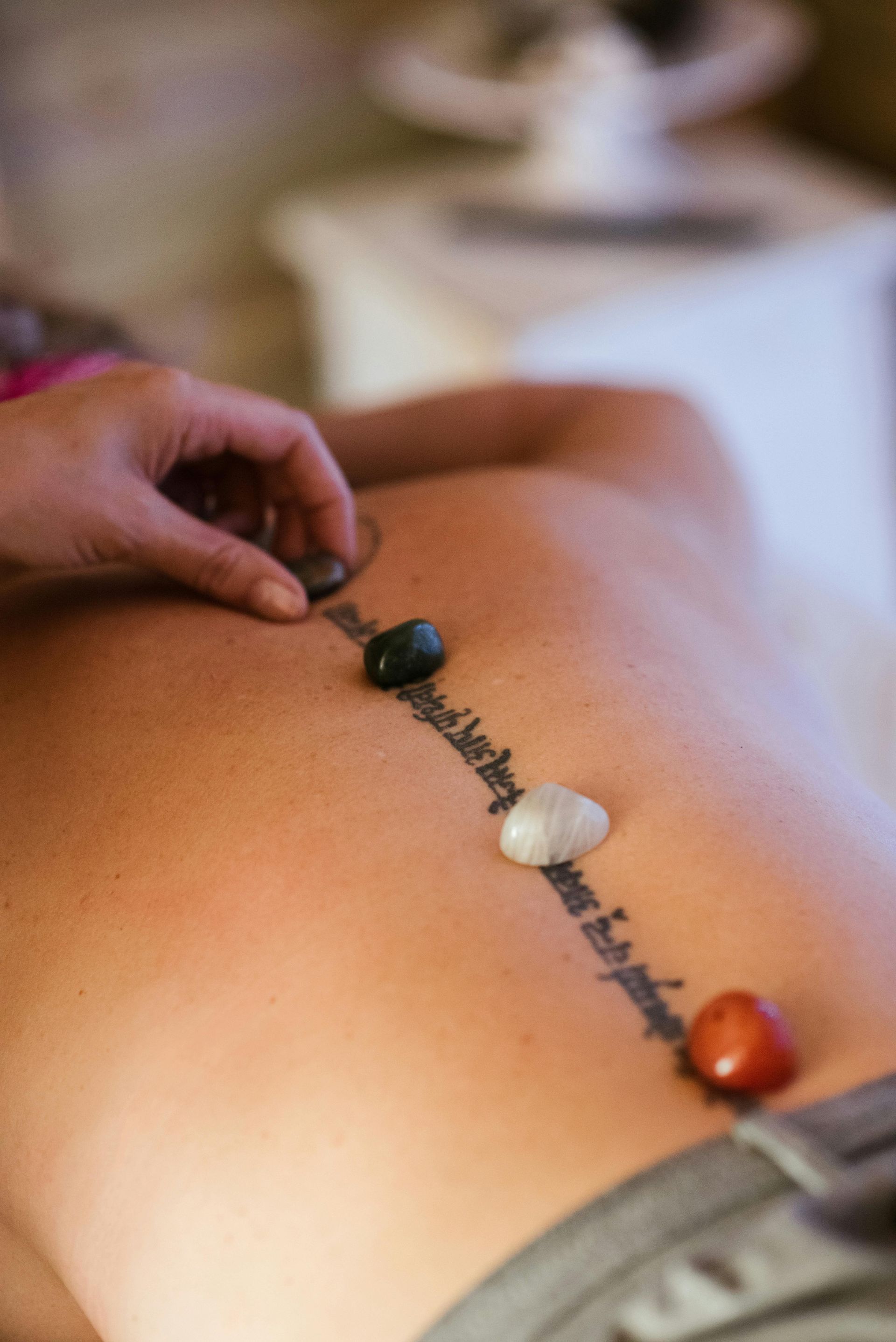 Hand placing stones along a spine with a tattoo. The stones are black, white, and red. The person is face down.