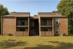 Two-story brown apartment building with balconies, set in a grassy area with trees in the background.