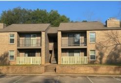 Two-story brick apartment building with balconies, beige siding, and a parking lot in front.