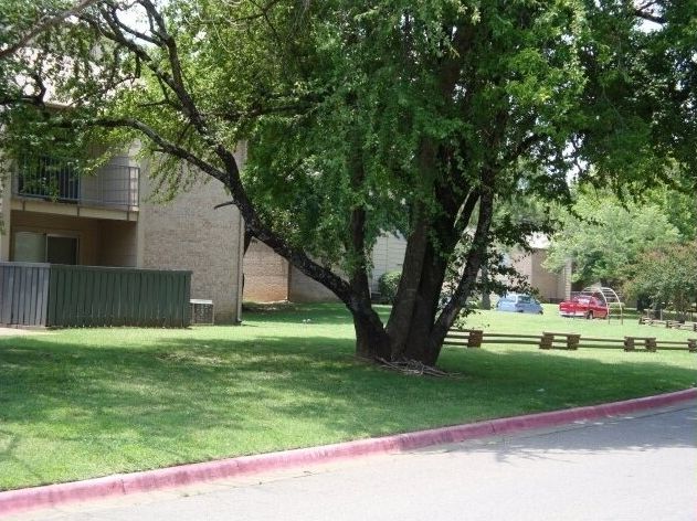 Green lawn with a large tree in front of an apartment building, picnic tables, and a red-curbed street.