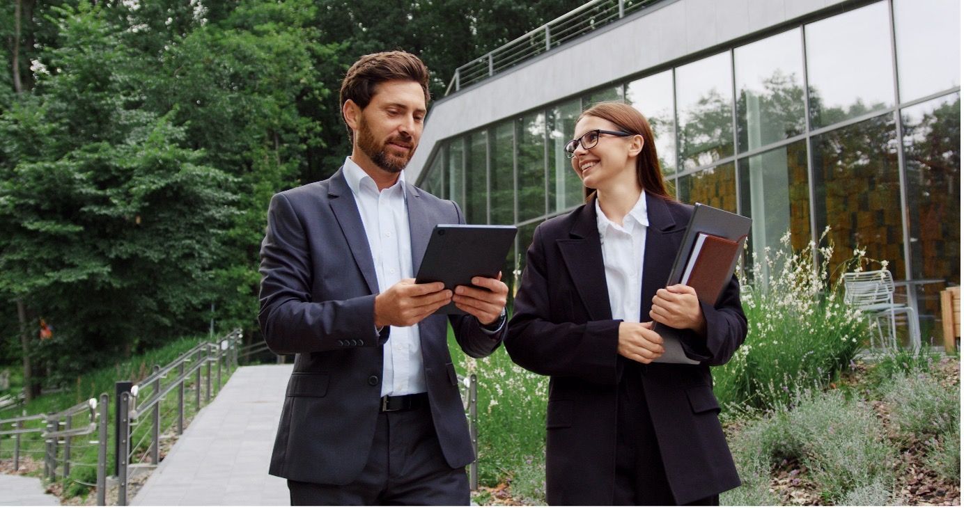 Two professionals in suits walk outdoors near a glass building, one holding a tablet and the other holding a folder.