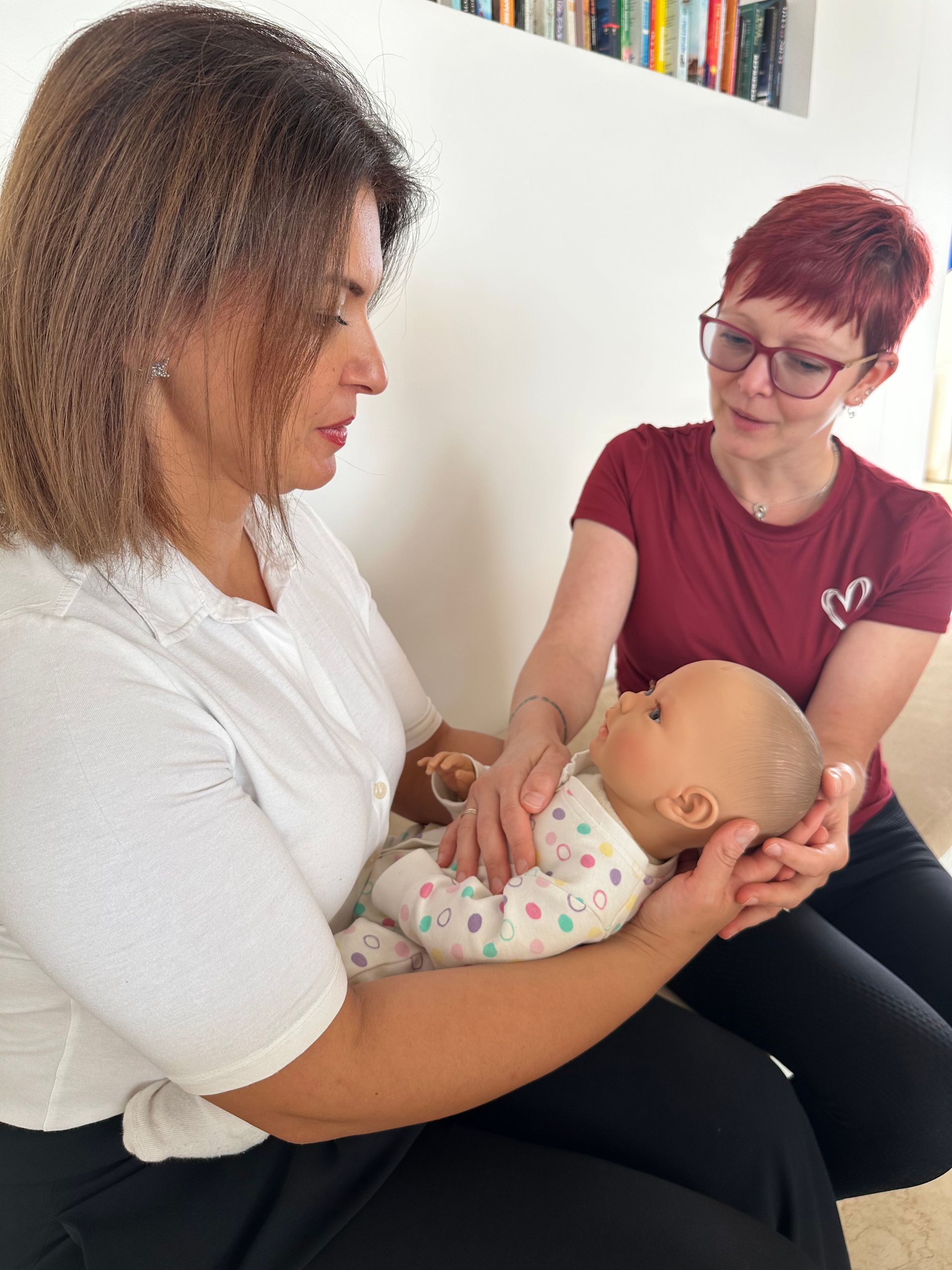 Zwei Personen demonstrieren die Säuglingspflege an einer Babypuppe; eine Person trägt ein weißes Hemd, die andere ein rotes.