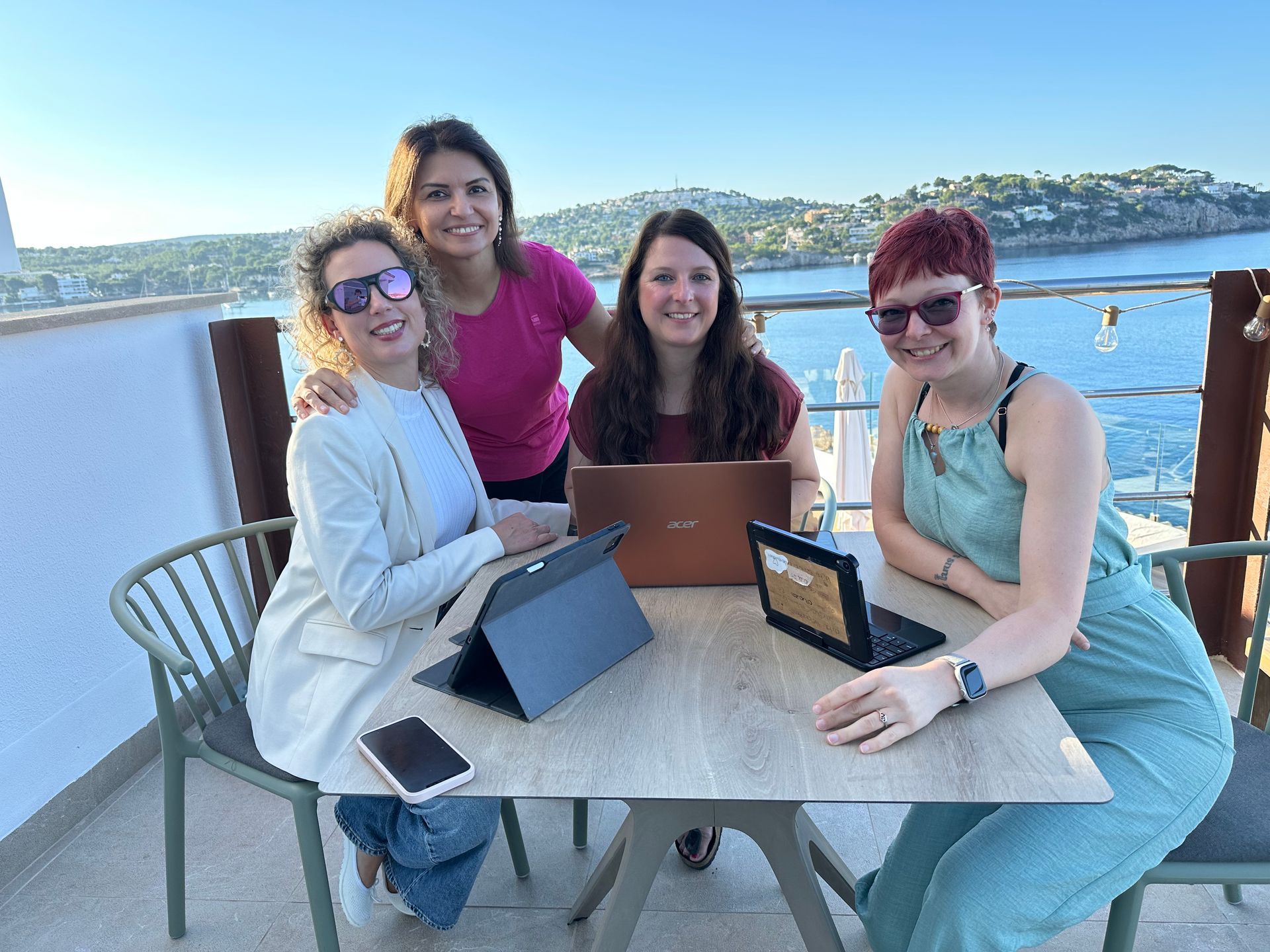 Vier Personen arbeiten an einem Tisch im Freien mit Laptops und Tablets und genießen den Blick auf die Küste.