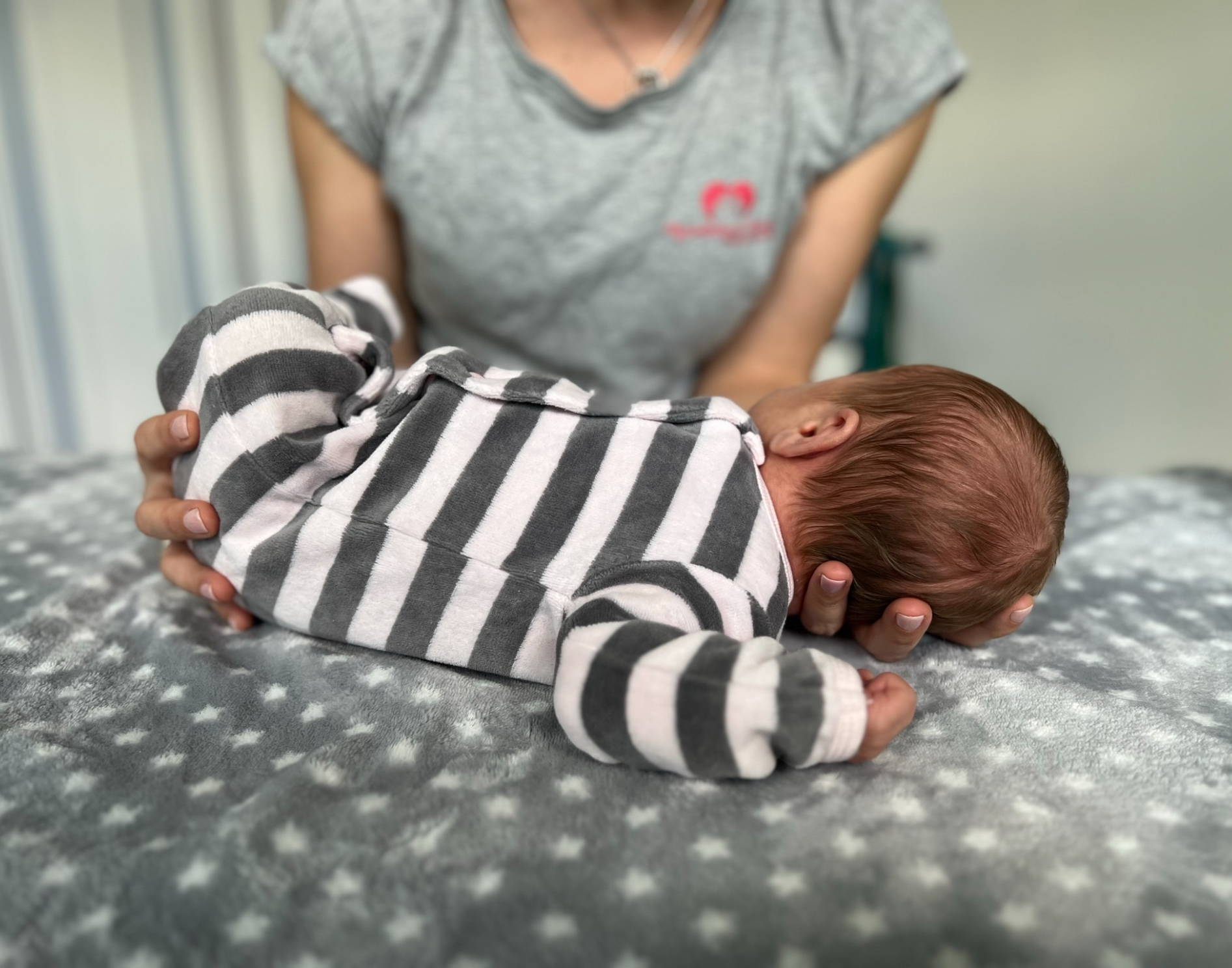 Baby auf einer grauen Decke. Es trägt einen gestreiften Strampler; dahinter steht eine Person in einem grauen Hemd.
