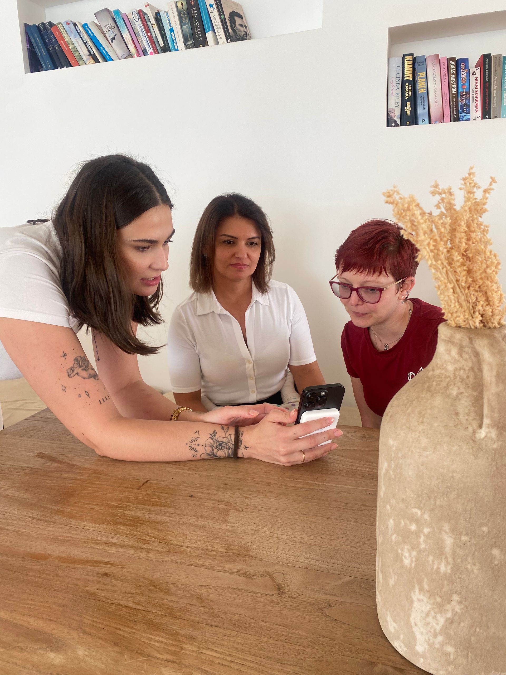 Drei Personen schauen auf ein Handy auf einem Holztisch. Die Frau links zeigt mit dem Finger, die anderen beiden schauen zu. 