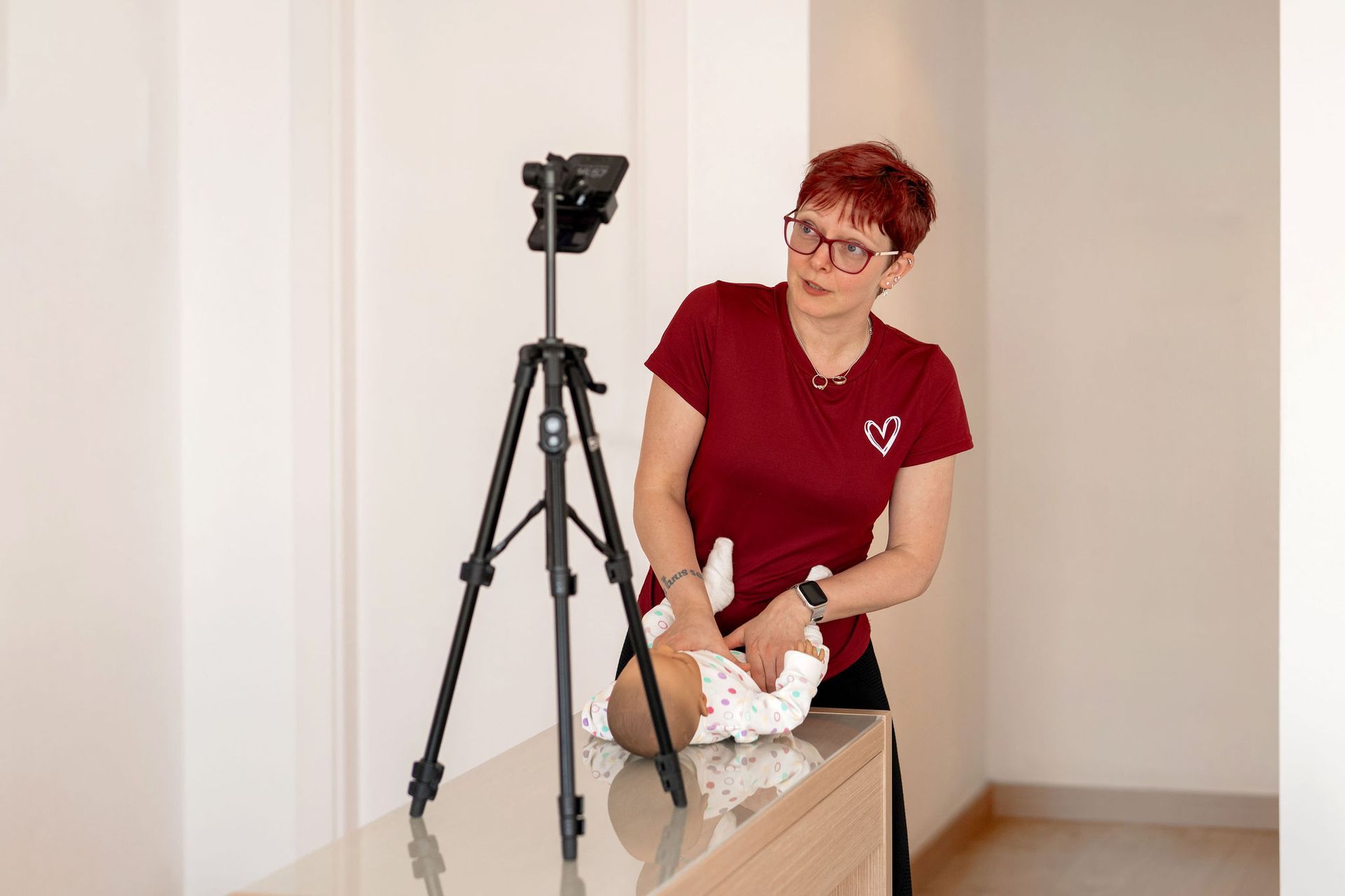 Eine Frau mit kurzen roten Haaren und Brille demonstriert die Verwendung einer Babypuppe, Stativ montierten Kamera.