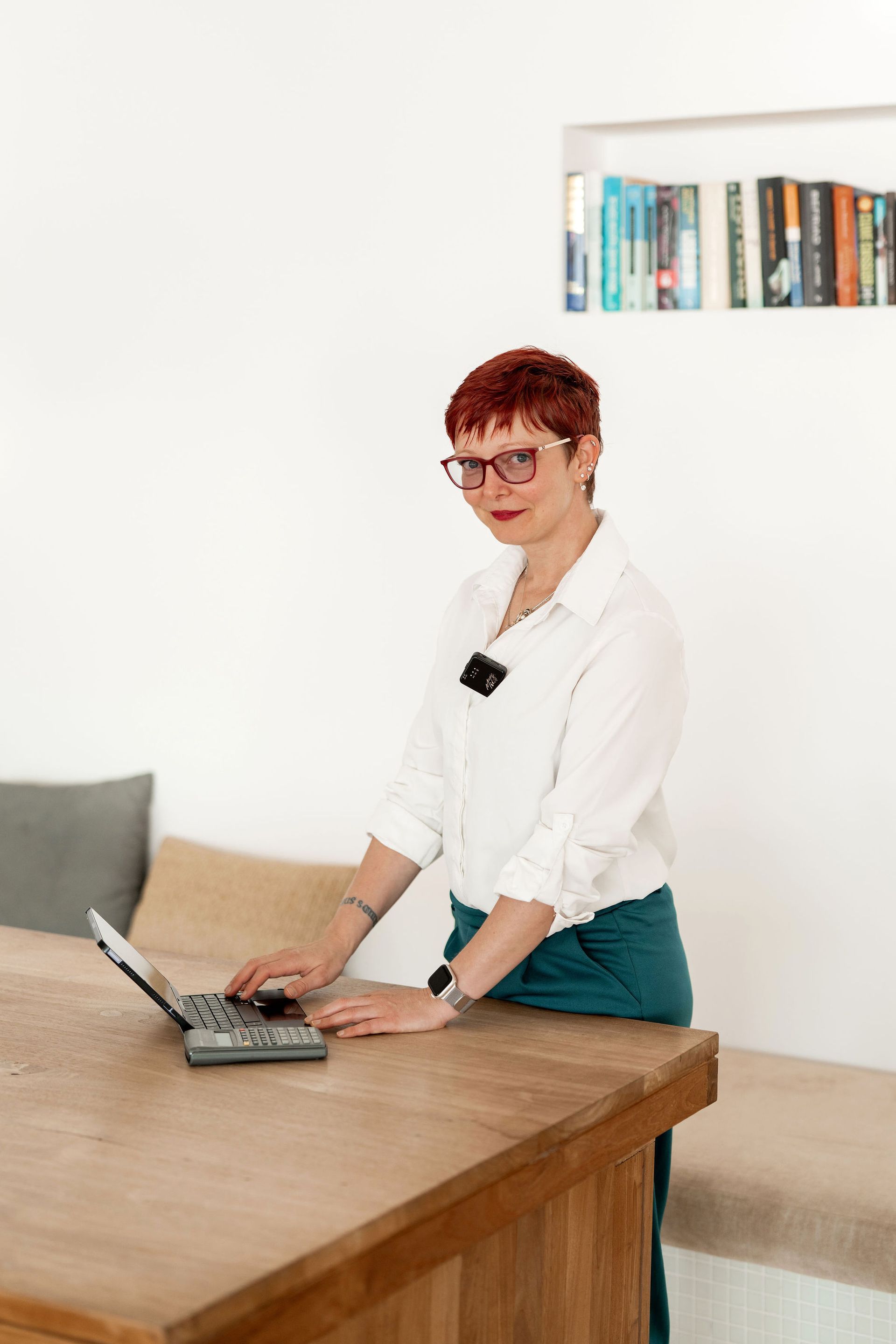 Frau mit kurzen roten Haaren und Brille arbeitet an einem Laptop an einem Holztisch. Weißes Hemd, türkisfarbene Hose.