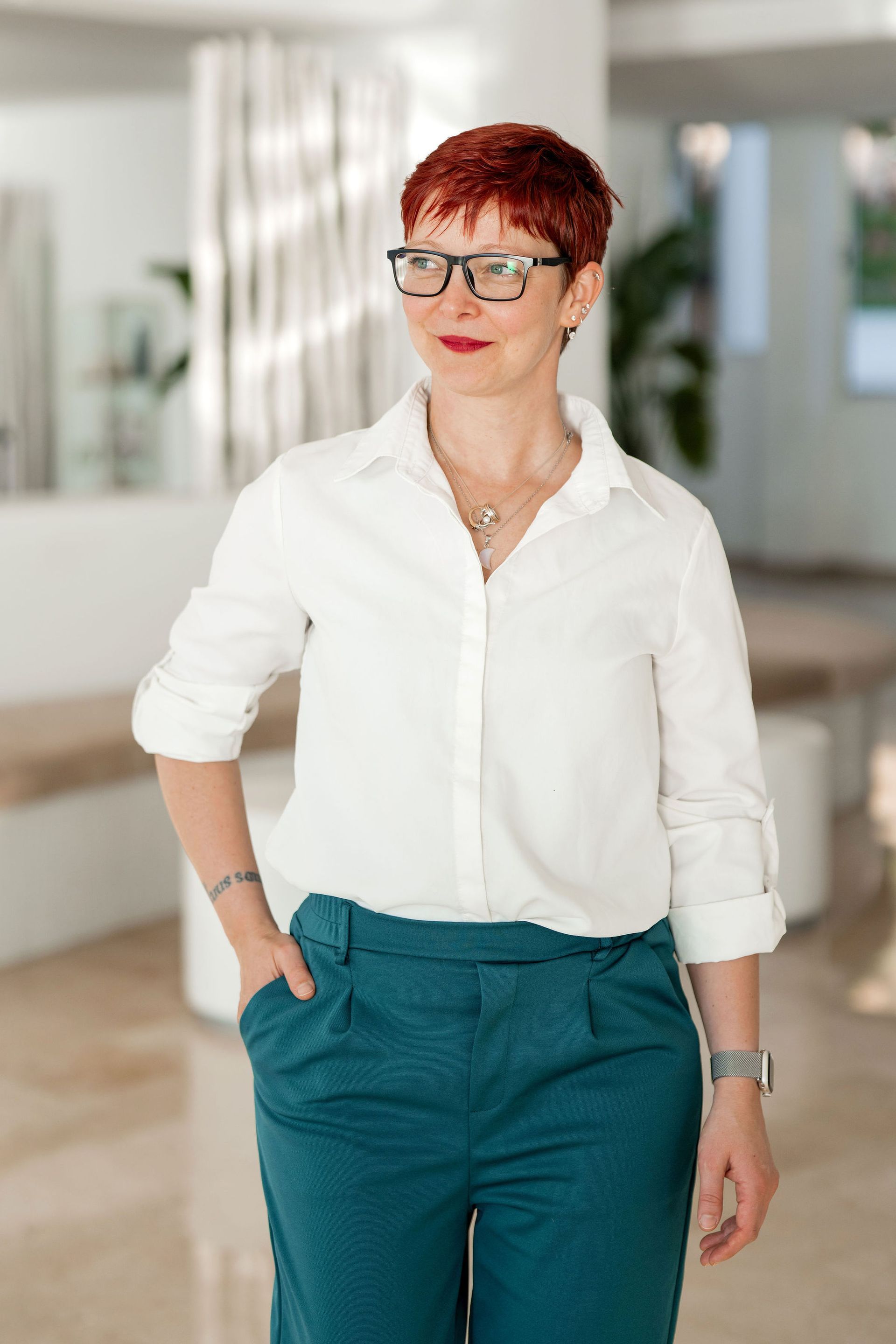 Frau mit kurzen roten Haaren, Brille, weißem Hemd, türkisfarbener Hose, Hand in der Tasche, blickt zur Seite.