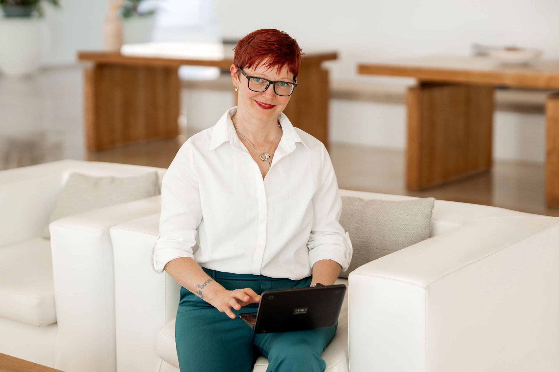 Frau mit roten Haaren und Brille arbeitet an einem Laptop auf einem weißen Sofa; helles, modernes Interieur.