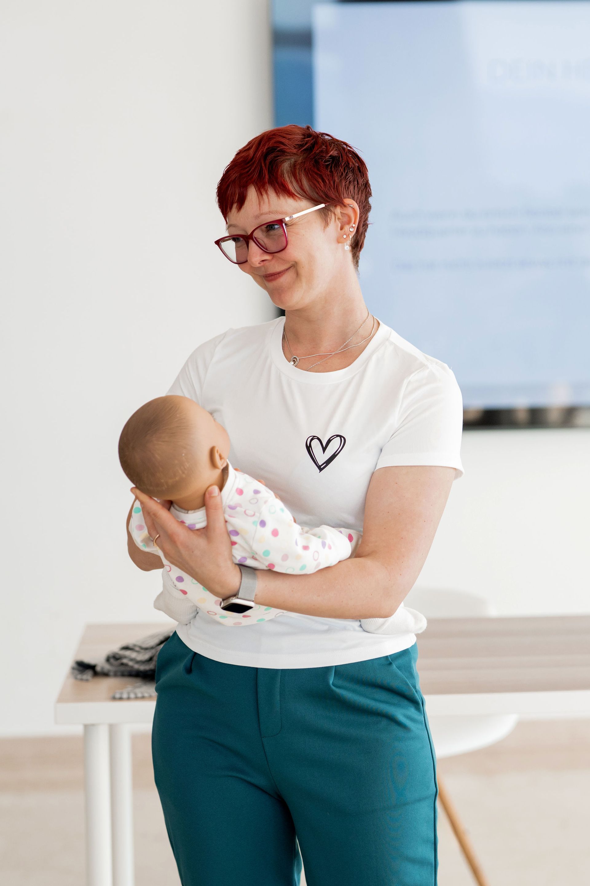 Frau mit Baby, die eine Brille und ein weißes Hemd mit Herzmuster trägt, in einem Innenraum.