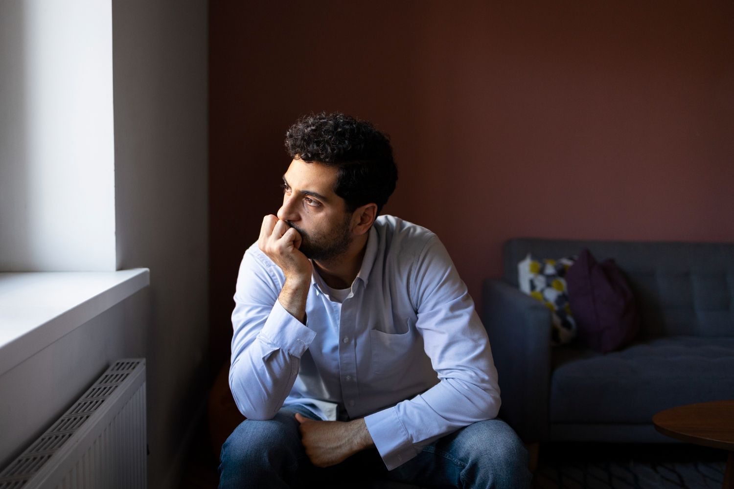 A man is sitting on the floor in front of a window looking out.
