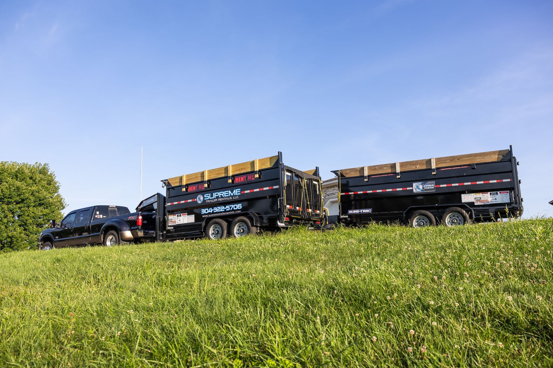 A black pickup truck hauling two black dump trailers on a grassy hillside under a blue sky.
