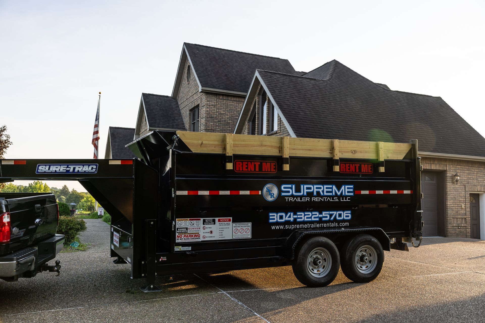 Black dump trailer hitched to a truck, parked in front of a brick house with a dark roof; the trailer has