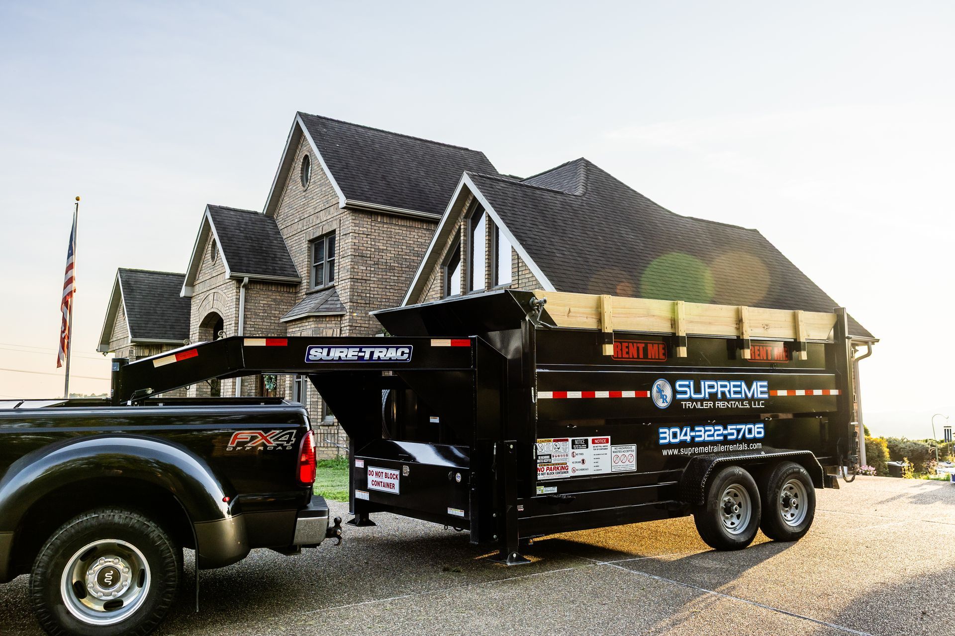 Black pickup truck towing a black Supreme Dumpsters trailer in front of a two-story brick house at sunset.