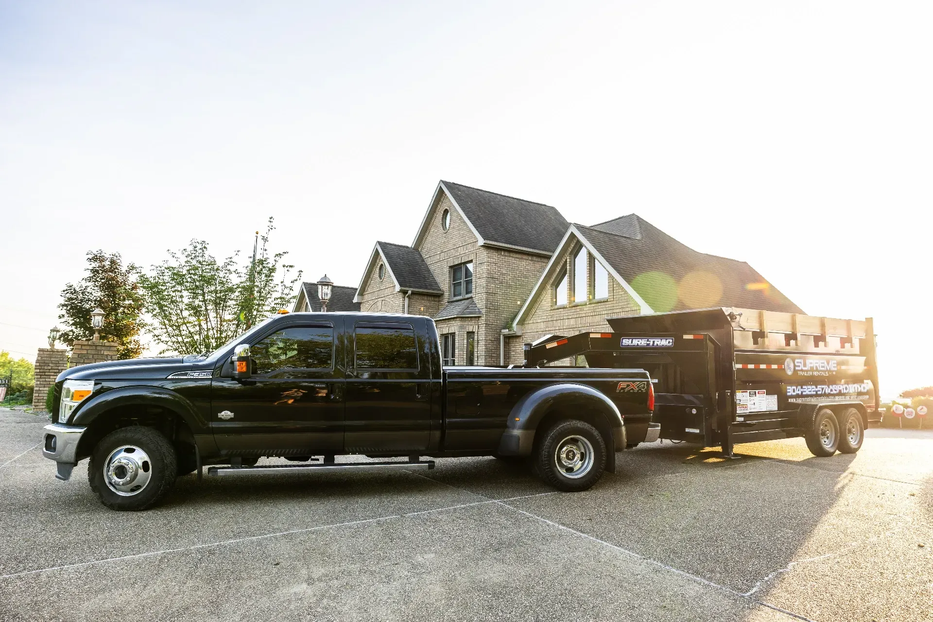 Black truck towing a trailer filled with materials, parked in front of a stone house under a bright sky.