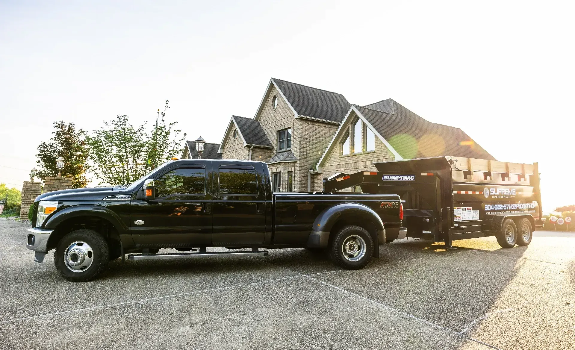 Black pickup truck towing a trailer loaded with items, parked on gravel in front of a large house.
