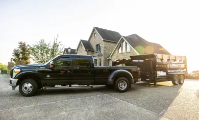 Black pickup truck towing a trailer loaded with wood, parked on gravel in front of a large house.