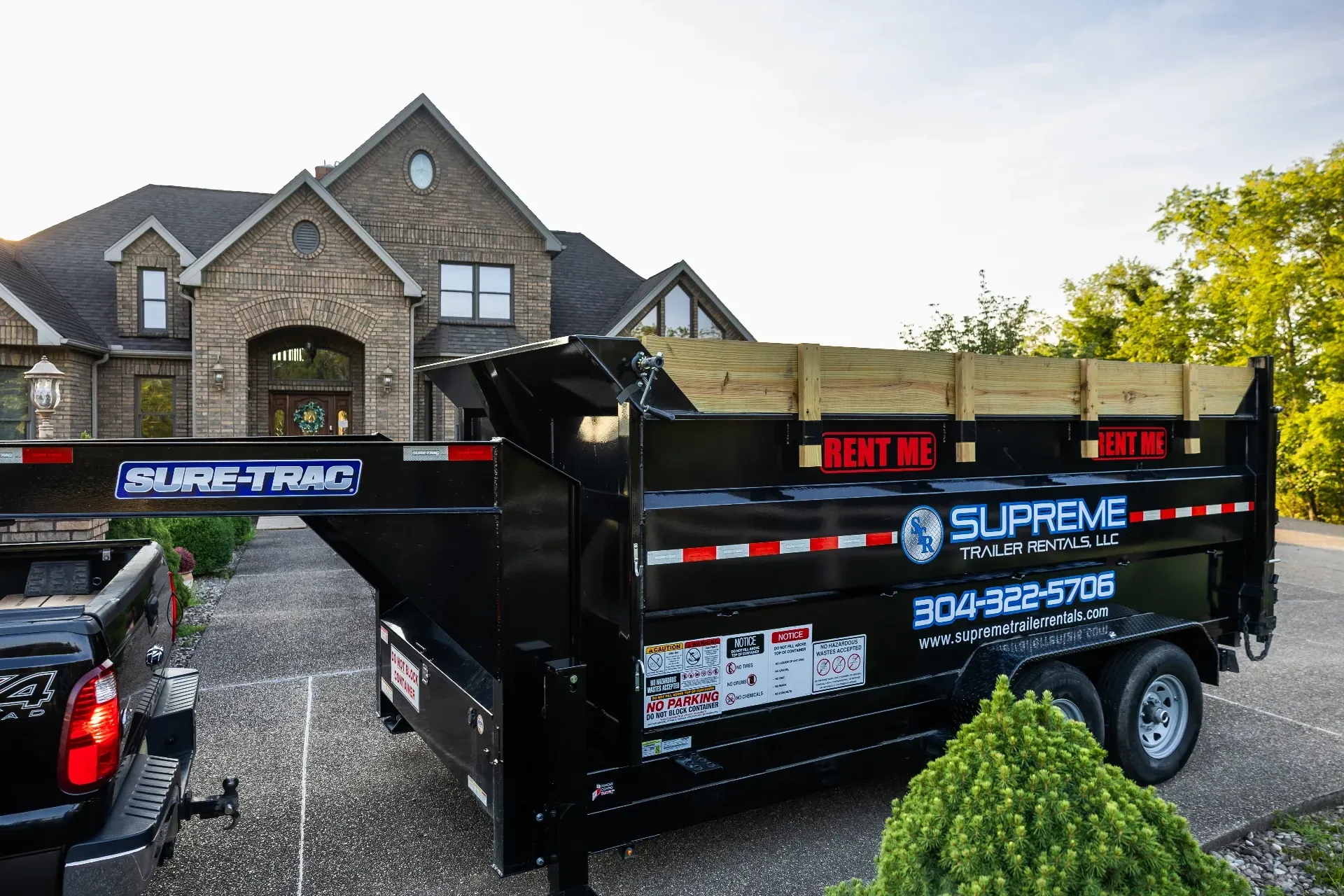 Black dump trailer parked in front of a large brick house, offering rentals.