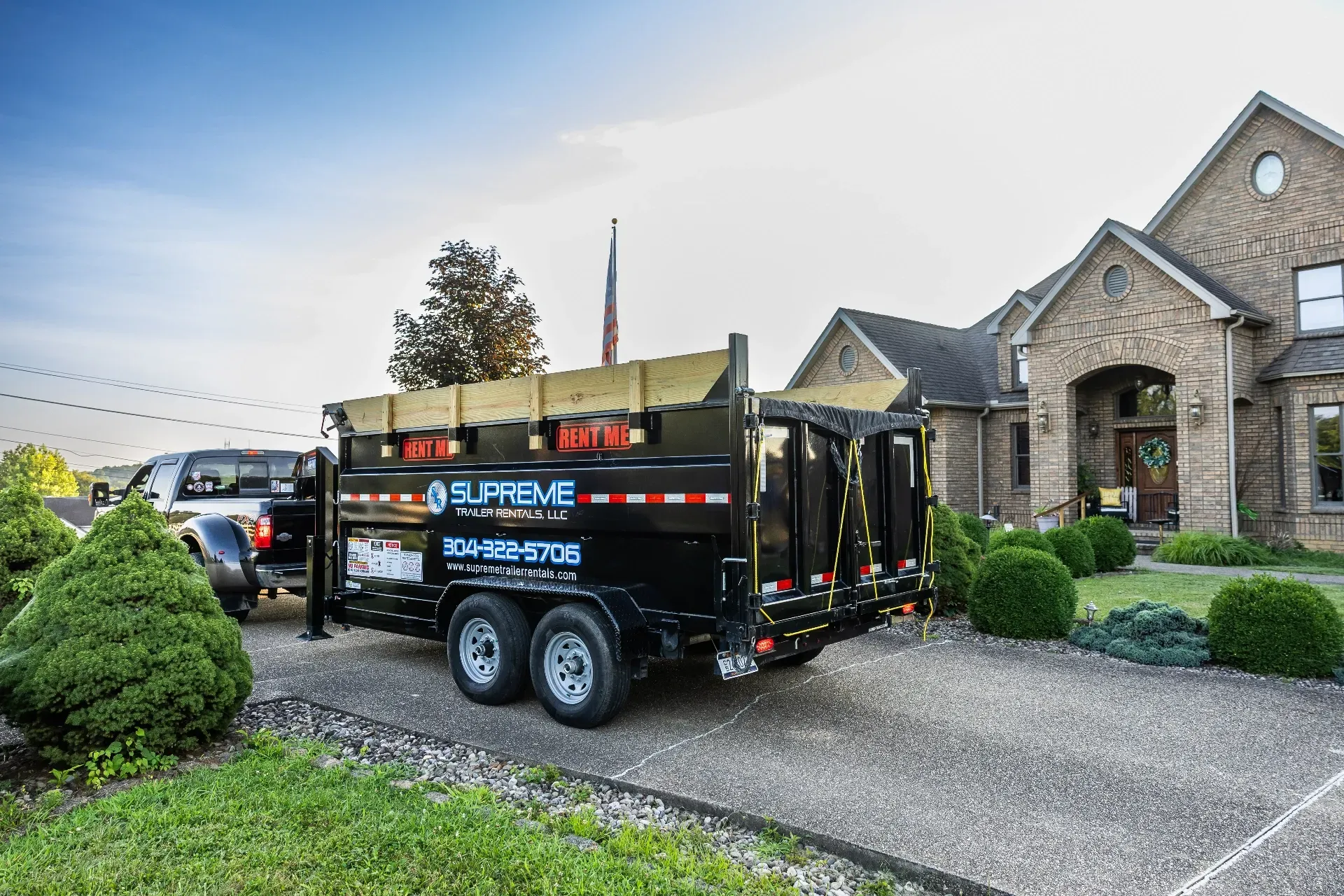 Black dump trailer attached to a truck parked on a gravel driveway in front of a brick house. The trailer has Supreme branding and a phone number.