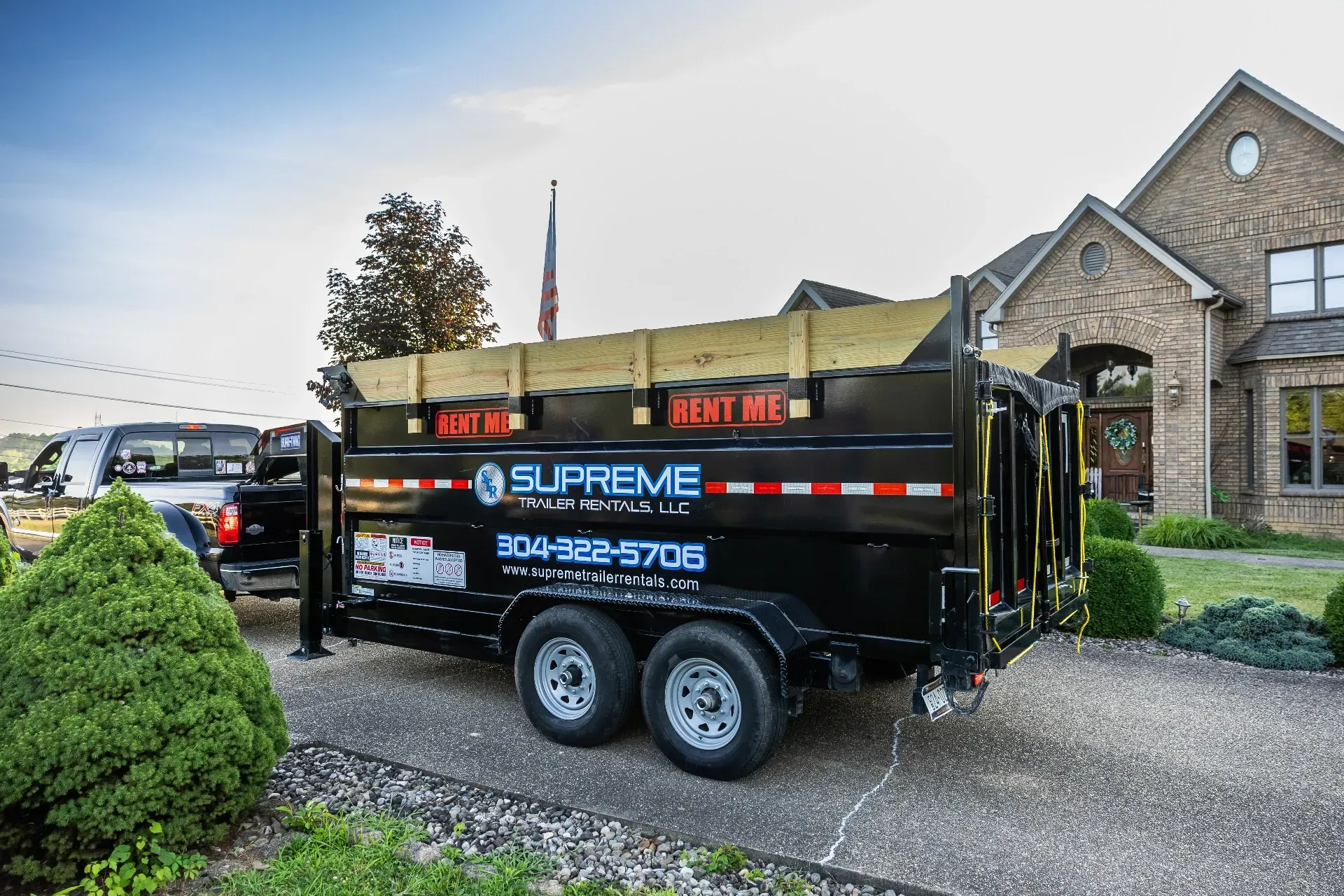 A black Supreme roll-off trailer is parked in a driveway next to a house.