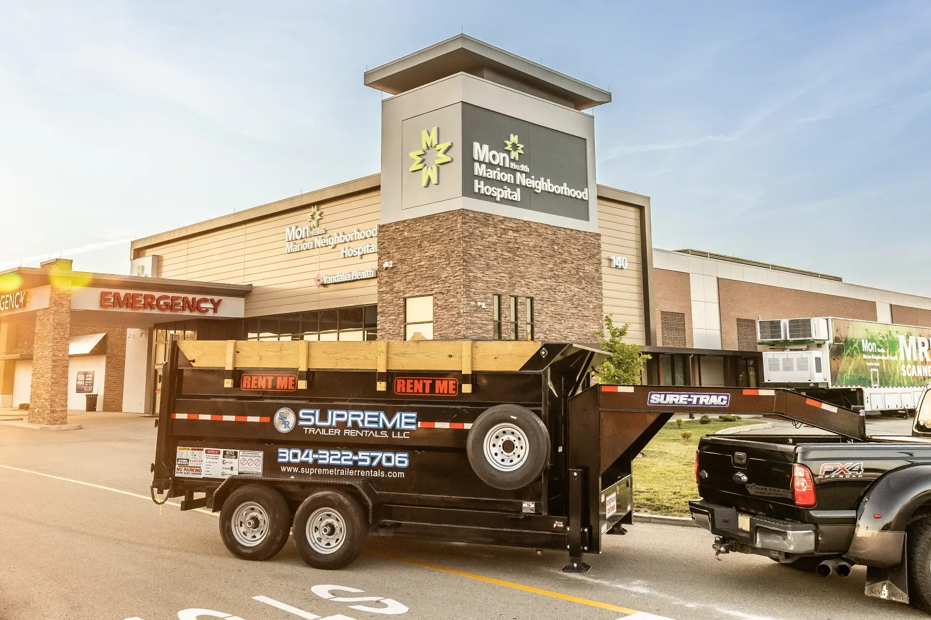 A black dump trailer is parked in front of a hospital. The trailer is attached to a black pickup truck.