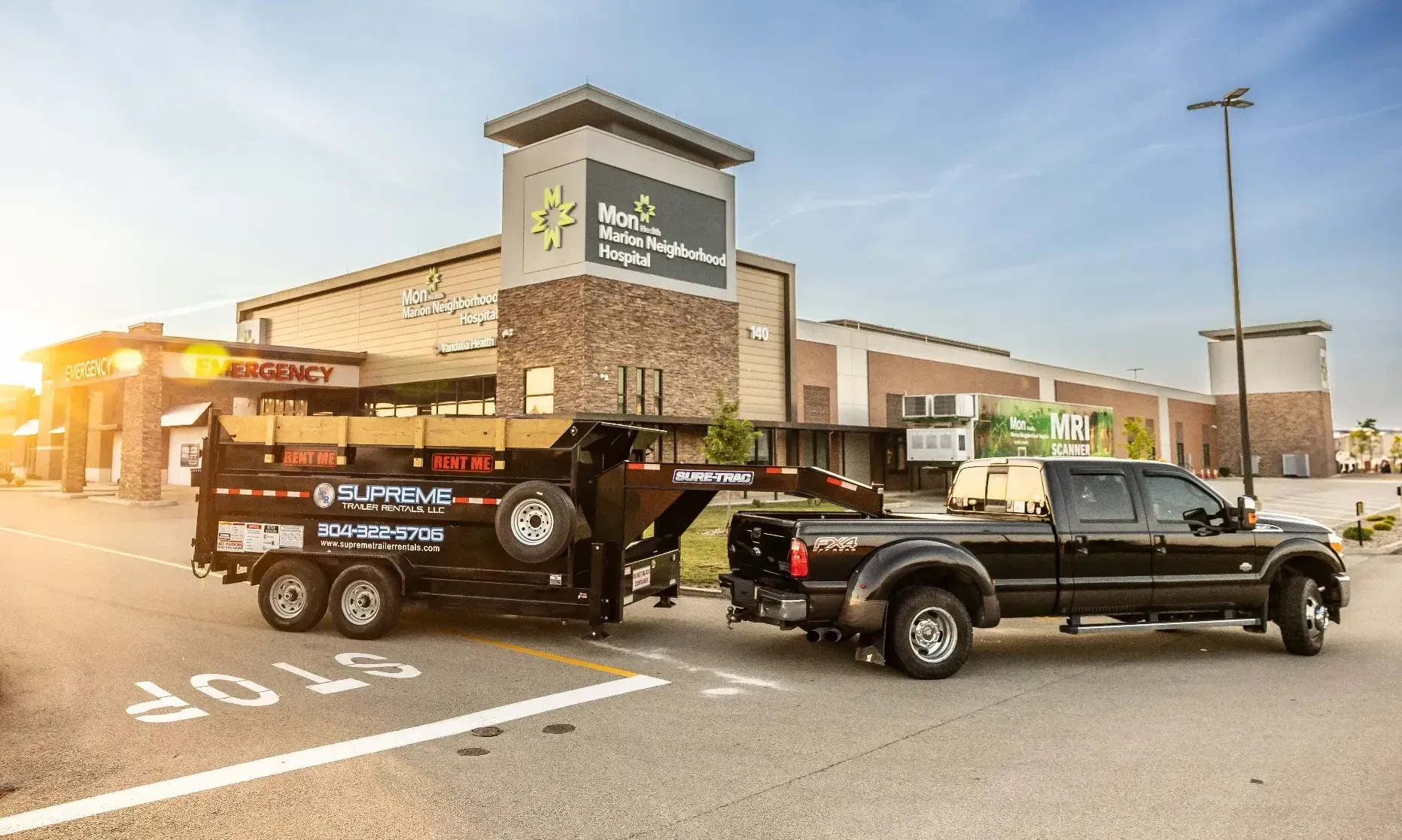 Black pickup truck towing a black trailer loaded with materials, parked in front of a shopping center with the sun setting.