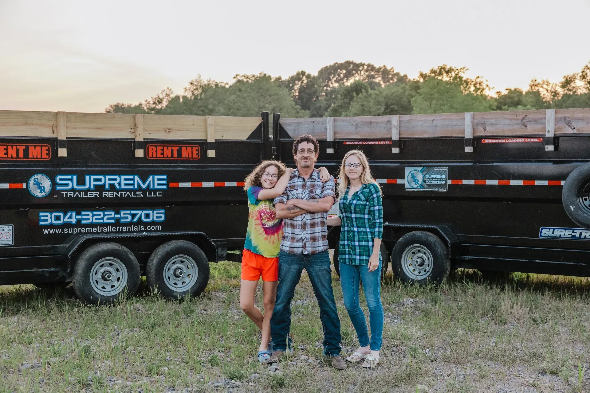 Three people posing in front of a black trailer. A man with crossed arms stands between two women, all smiling. The setting is outdoors.