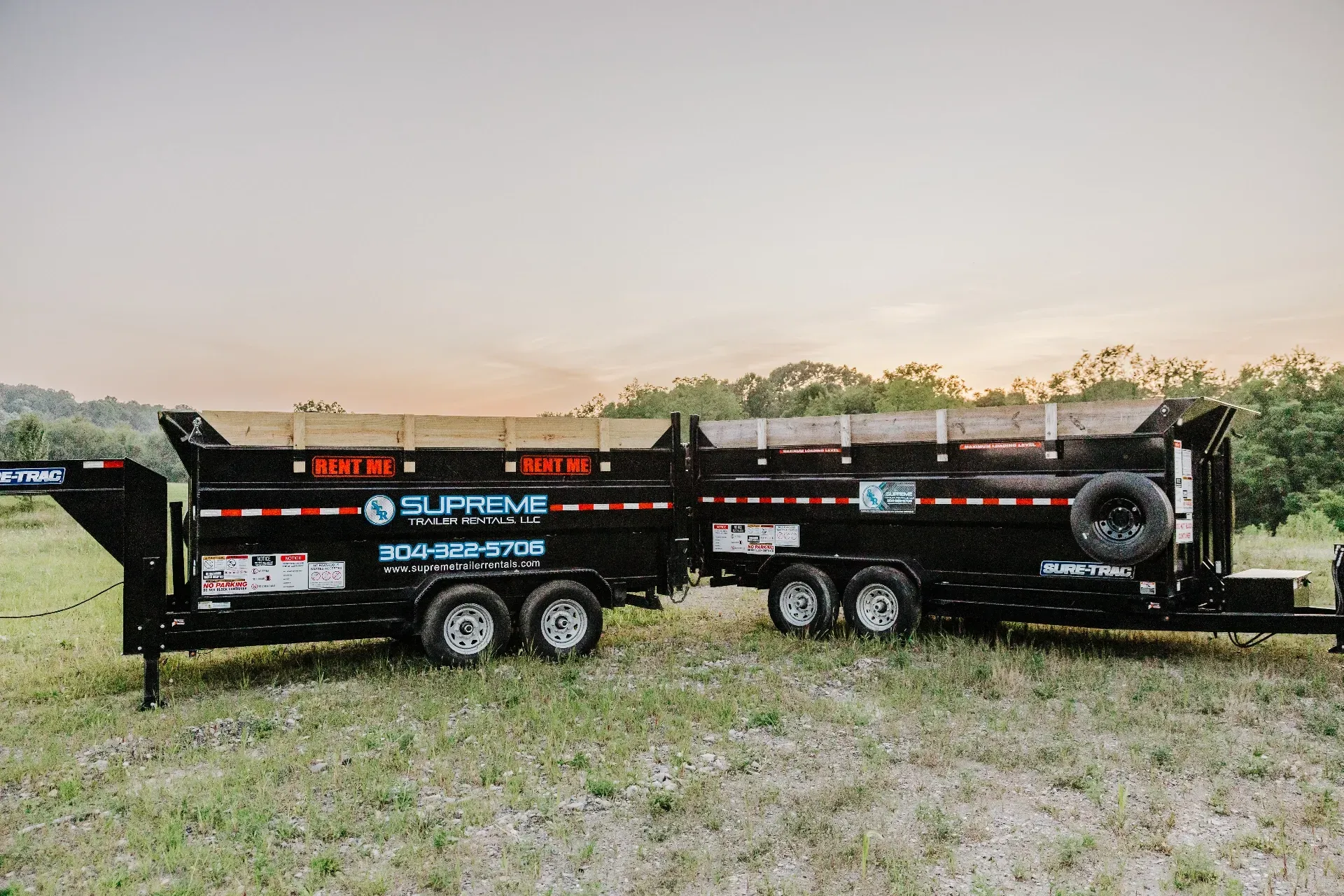 Two black dump trailers side-by-side on a grassy field, with a sunrise or sunset in the background.