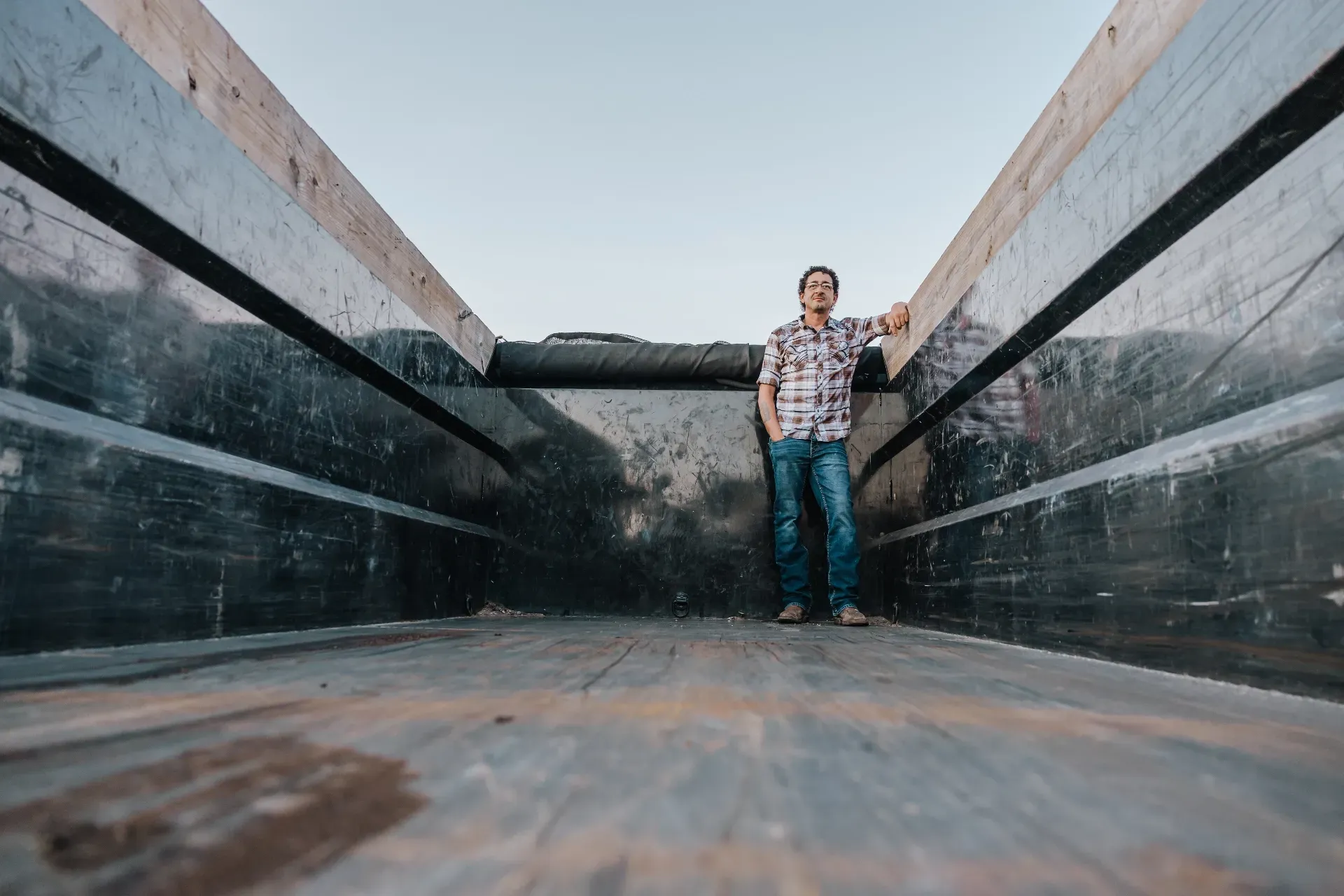 Man in plaid shirt and jeans stands in a truck bed, looking forward. Sky is visible overhead.