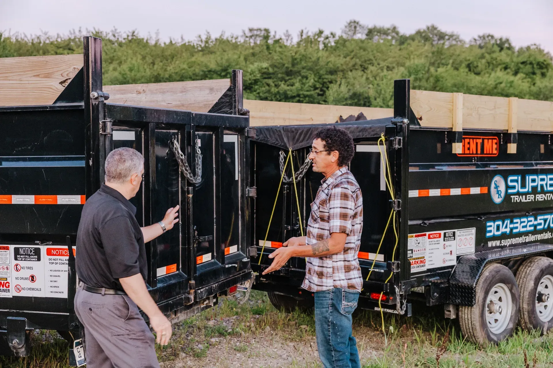 Two men inspecting the open back doors of two black dump trailers in a grassy outdoor setting.