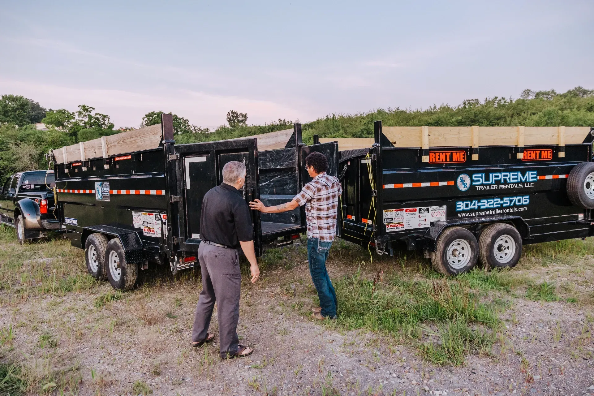 Two men inspecting dump trailers. The trailers are black with blue and white logos. They are outdoors.
