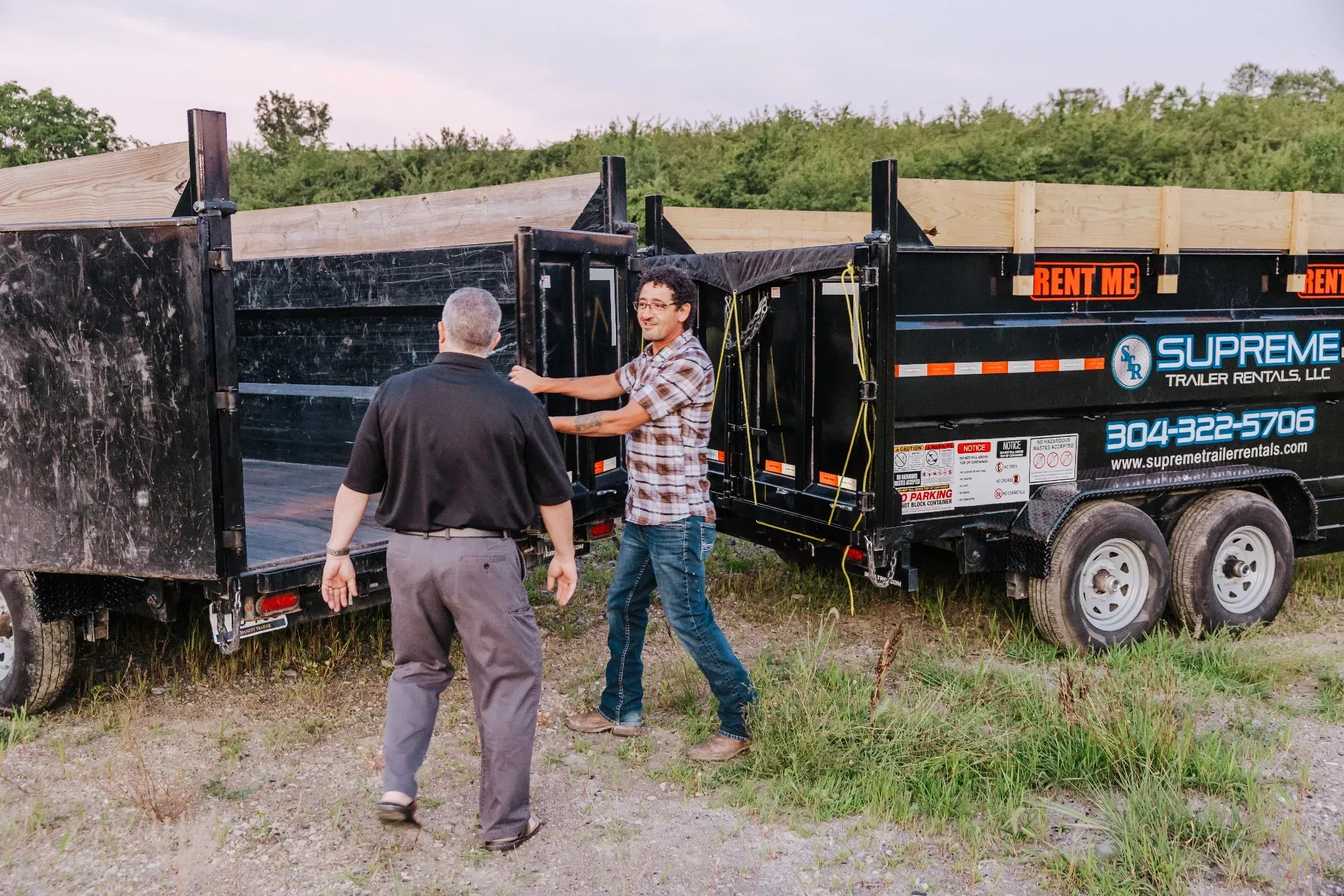 Two men beside dump trailers. One man opens a trailer door while the other watches. Outdoor setting, daytime.