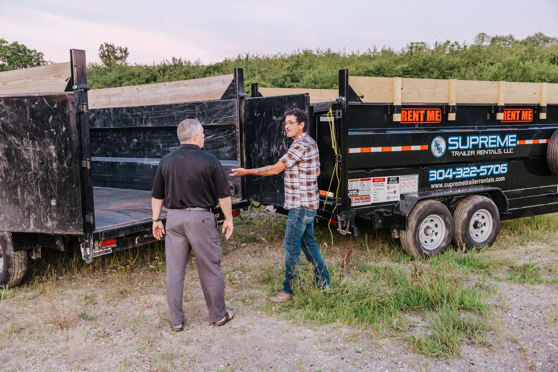 Two men near black Supreme Rentals dump trailers outdoors. One man points towards the trailer while the other faces him.